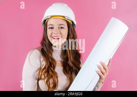 Portrait of a happy female architect builder in the hard hat holding layout plan on the pink background. Stock Photo