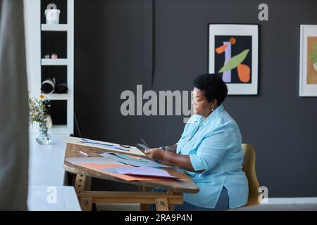 Side view of creative senior black woman in casualwear cutting paper with scissors while working over creation of new masterpiece in studio Stock Photo