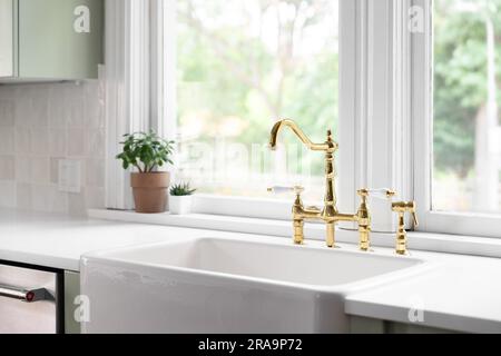 A kitchen faucet in a cozy green kitchen with a gold bridge faucet, white apron sink, tiled backsplash, and white marble countertop. Stock Photo