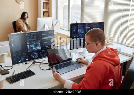 Side view of young female cyber security engineer using smartphone while sitting by workplace in front of three computers with data on screens Stock Photo