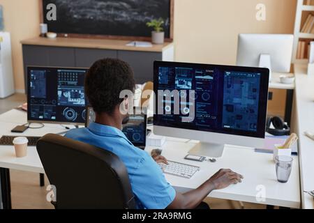 Rear view of young African American businessman sitting by workplace in front of desktop computers with graphic data on screens in office Stock Photo