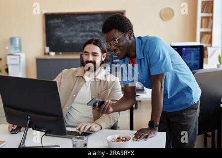 Two young smiling male IT managers looking at data on computer screen while one of them explaining data to colleague or consulting him Stock Photo