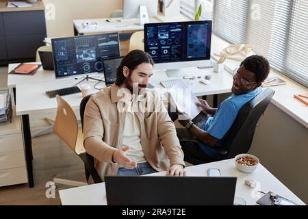 Young businessman showing data on computer screen to smiling African American colleague while explaining him results of analyzing it Stock Photo
