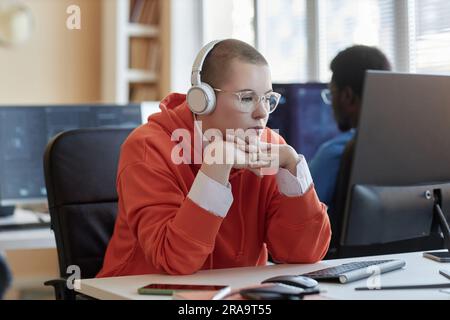Young IT support manager or cyber security engineer in headphones sitting in front of computer and looking at screen against colleague Stock Photo