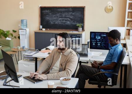 Young serious cyber security manager typing on computer keyboard while sitting by workplace and working with online data against colleague Stock Photo