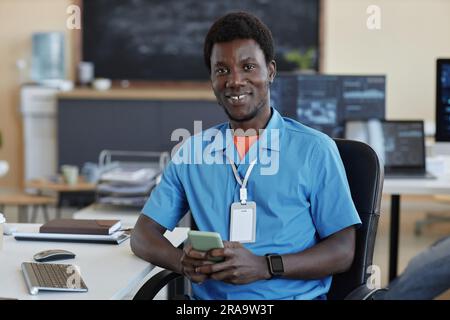 Young successful African American programmer or cyber security manager texting in mobile phone and looking at camera while sitting by desk Stock Photo