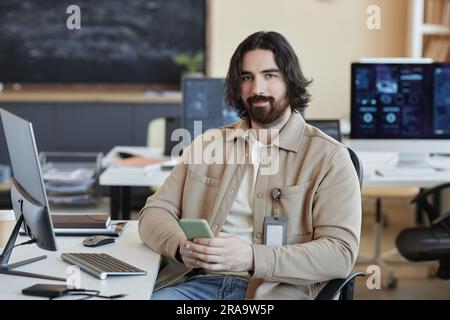 Young bearded IT engineer with smartphone texting and looking at camera while sitting by workplace in office and working over new software Stock Photo