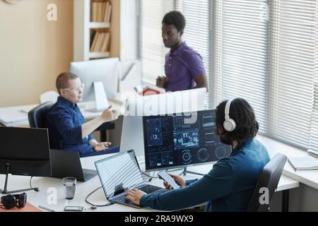 Young worker of cyber security center sitting in front of computers with graphic data and using smartphone while his colleagues having discussion Stock Photo
