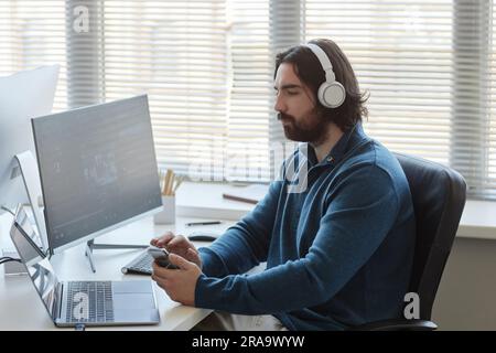 Young man in headphones using mobile phone during work with data while sitting by workplace in front of laptop and computer screen Stock Photo