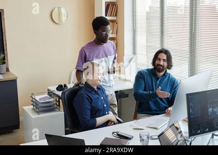 Confident smiling businessman pointing at computer screen during discussion of online data with two young intercultural colleagues Stock Photo