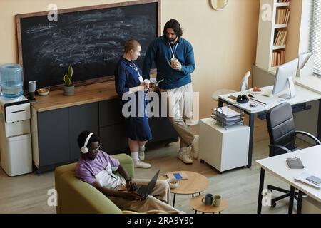 Two young managers discussing video in smartphone and having coffee at break while standing in front of coworker in headphones using laptop Stock Photo