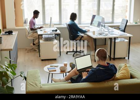Rear view of young female IT manager with laptop sitting on couch in office and networking in front of two intercultural male colleagues Stock Photo