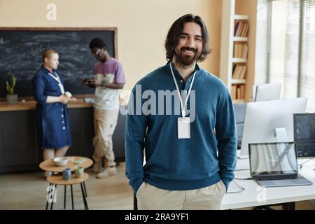 Young successful businessman in quiet luxury attire looking at camera with smile while standing against two intercultural colleagues having chat Stock Photo