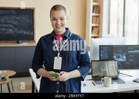 Young smiling businesswoman using mobile phone and looking at camera while standing by workplace with group of desktop computers Stock Photo