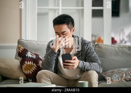 young asian sitting on couch at home covering eyes with hand feeling tired after looking at cellphone for long time Stock Photo