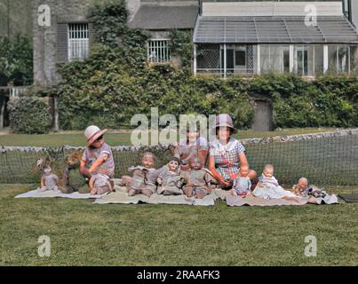 Three girls sitting in a garden with an array of dolls and one teddy bear.  There is a low tennis net behind them. Stock Photo