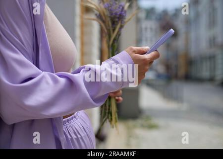 Stylish young woman in purple clothes using a smart phone in purple cover. Unrecognizable female person standing in the city street with a cellphone i Stock Photo