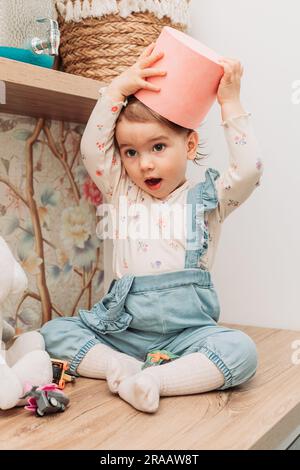 Little baby girl sitting on the table putting paper bucket on her head. Stock Photo