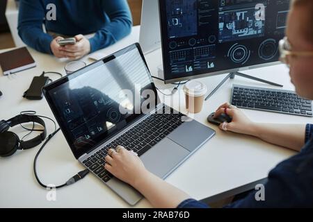Young female analyst or IT manager using laptop while sitting by workplace in front of computer monitor with graphic data Stock Photo