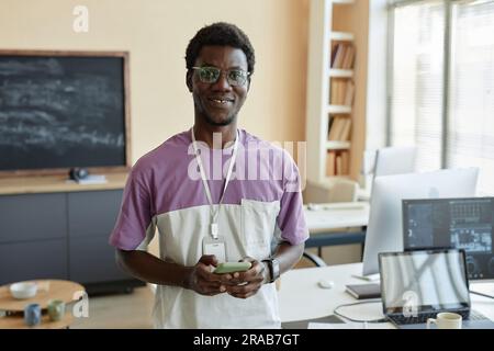 Young smiling manager in eyeglasses and casualwear using mobile phone and looking at camera while standing against workplace with computers Stock Photo