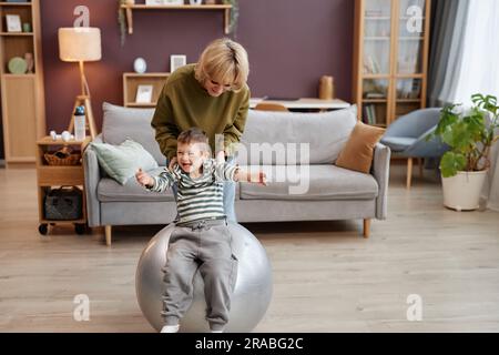 Full length portrait of cute little boy with down syndrome having fun jumping on fitness ball with caring mother assisting, copy space Stock Photo