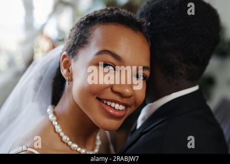 Close up portrait of black young woman smiling at camera laying head on grooms shoulder in wedding ceremony Stock Photo