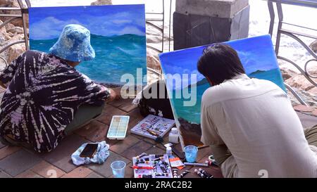 Two young men paint seascapes on canvases as they sit beside the sea in Pattaya, Thailand; painting painters art artistic theme Stock Photo