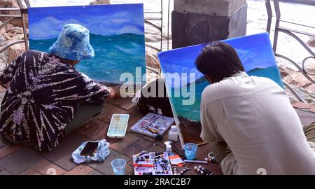 Two young men paint seascapes on canvases as they sit beside the sea in Pattaya, Thailand; painting painters art artistic theme Stock Photo