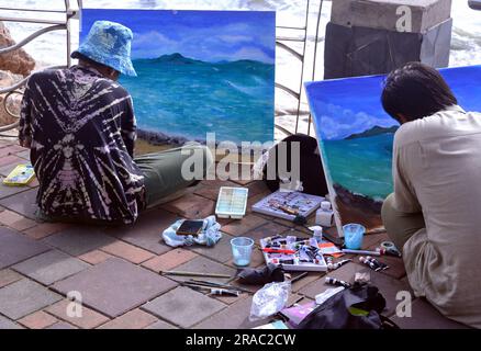 Two young men paint seascapes on canvases as they sit beside the sea in Pattaya, Thailand; painting painters art artistic theme Stock Photo