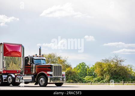 Industrial American old style day cab burgundy big rig semi truck tractor with high chrome pipes transporting cargo in bulk semi trailer with red spoi Stock Photo