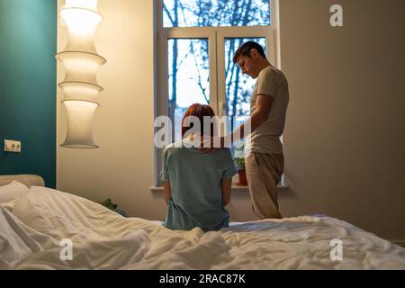 Family support. Sad woman sits on bed near standing caring loving man husband touching wife shoulder Stock Photo