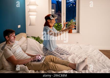 Focused boyfriend sits on bed with laptop near girlfriend playing virtual game wearing VR 3D glasses Stock Photo
