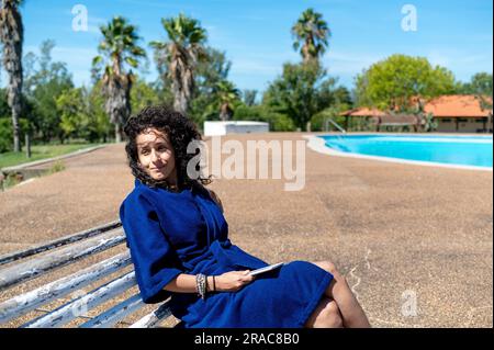 Woman relaxing sitting on a bench outdoors near swimming pool. Stock Photo