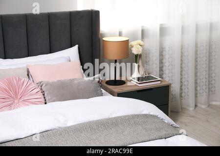 Comfortable bed with cushions, lamp and different decor on bedside table in room. Stylish interior Stock Photo