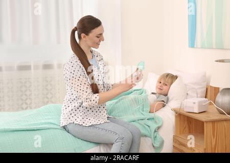 Mother holding nebulizer for inhalation and her son lying on bed at home Stock Photo