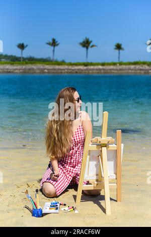 Young artist woman sitting at the beach with a jar with paint brushes and easel close to sea and palms in summer Stock Photo