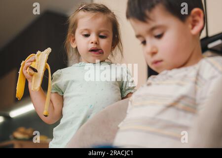 Cute little girl in casual clothes sitting on sofa and eating banana while spending time in cozy room Stock Photo