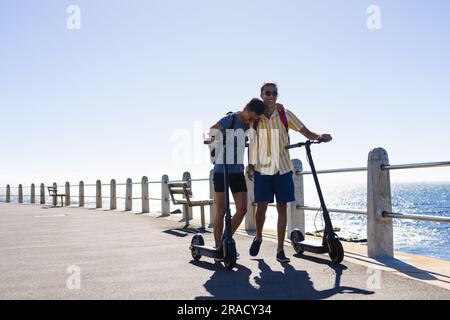 Happy biracial gay male couple walking with scooters on promenade by the sea, copy space Stock Photo