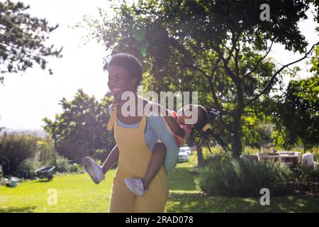 African american cheerful mother piggybacking daughter while standing against trees in park Stock Photo