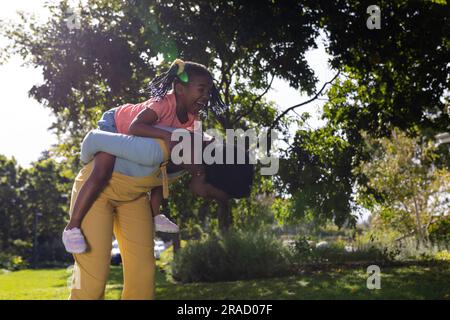 African american playful mother piggybacking daughter while bending against trees in park Stock Photo