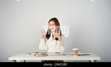 A cheerful and smiling young Asian businesswoman or female office worker sits at her desk and shows her thumb up. isolated white background Stock Photo