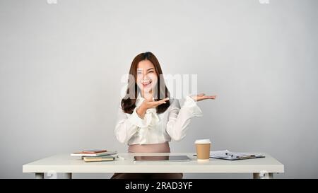 A beautiful and smiling Asian businesswoman or female office worker sits at her desk and opens her palms to the empty space. recommend, advice, giving Stock Photo