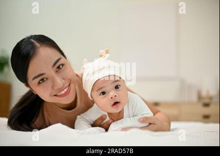 Sweet family moment. Young Asian mom lay down with her baby, smiling to camera. showing affection to her baby. Stock Photo