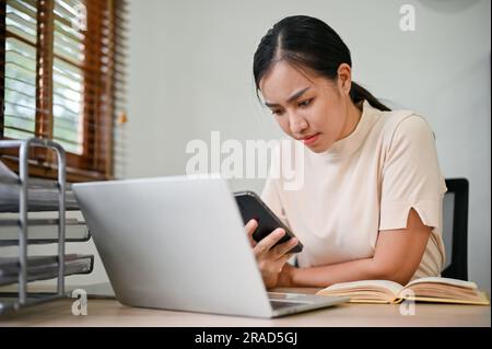 Young stressful woman working from home with smartphone and laptop computer. Stock Photo