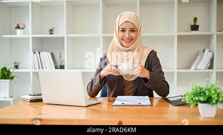 A portrait of a beautiful and successful Asian Muslim businesswoman or female boss in hijab, smiling and looking at the camera while sitting at her de Stock Photo