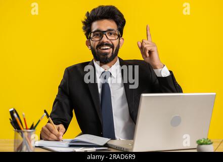 Thoughtful journalist indian businessman making notes, writing down thoughts with pen into notepad notebook diary, to do list, good idea at office workplace desk. Hindu man in suit working on laptop Stock Photo