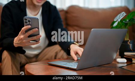 Relaxing young Asian man sitting oh their sofa and holding smartphone in his hand in free time. Stock Photo