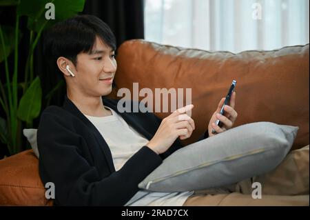Relaxing young Asian man lay down comfortably on the sofa, using smartphone in his free time Stock Photo