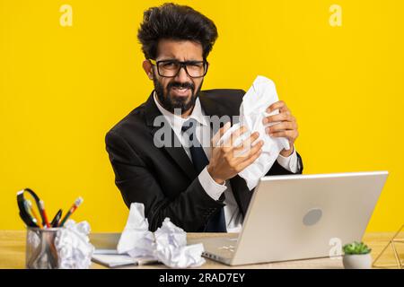 Angry furious indian man working at office throwing crumpled paper, having nervous breakdown at work, migaine, headache, stress management, mental distress problems, losing temper, reaction on failure Stock Photo