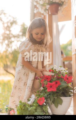 little girl with blonde hair is watering flowers on the veranda in summer at sunset. High quality photo Stock Photo